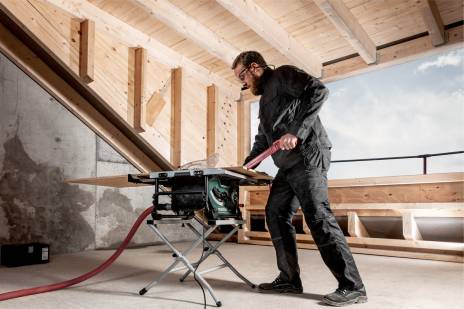Worker in a loft conversion using a Metabo table saw on a stand, pushing a plywood panel through the saw, with a dust extraction hose attached.
