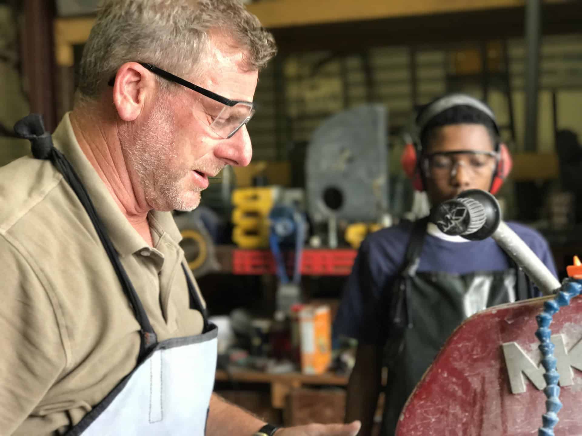 Middle-aged tradesperson wearing safety glasses and a work apron operating a machine, while an apprentice in safety glasses and earmuffs observes closely in a workshop setting, safety lesson